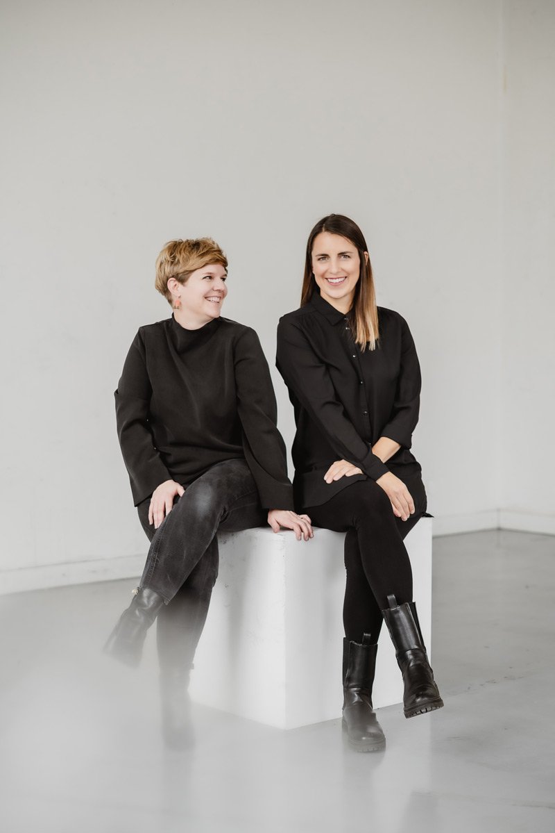 Foto der Geschäftsführerinnen Isabelle Possehl und Janina Schneider: Beide sitzen lächelnd nebeneinander auf einem weißen Würfel vor einer weißen Wand und wirken entspannt und professionell.