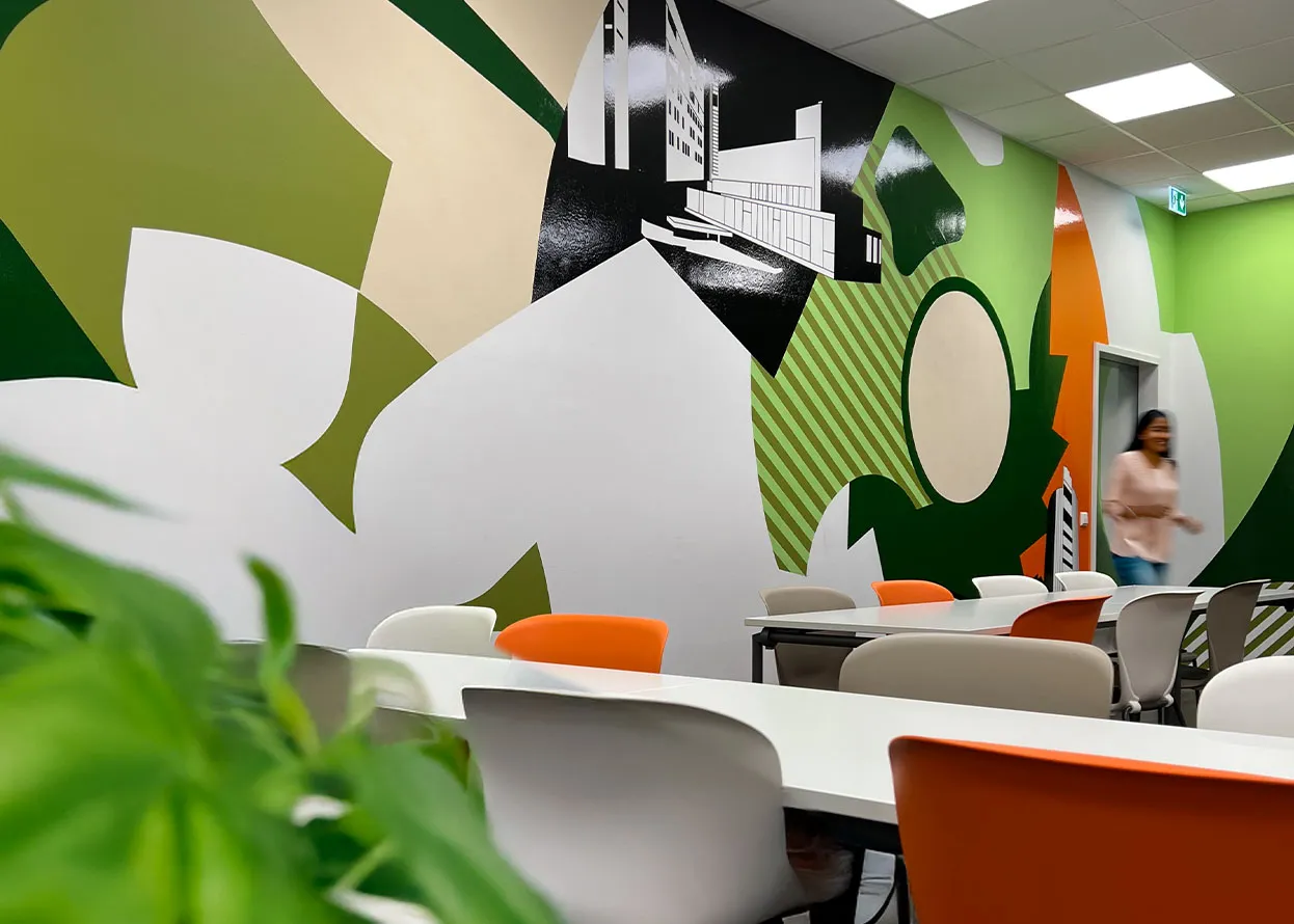 Besprechungsraum mit langem weißem Tisch, orangefarbenen und weißen Stühlen. Die Wand ist mit einem Tape-Art-Werk aus abstrakten Formen in Grün-, Orange- und Weißtönen gestaltet. Links im Bild ist eine verschwommene Person zu erkennen.